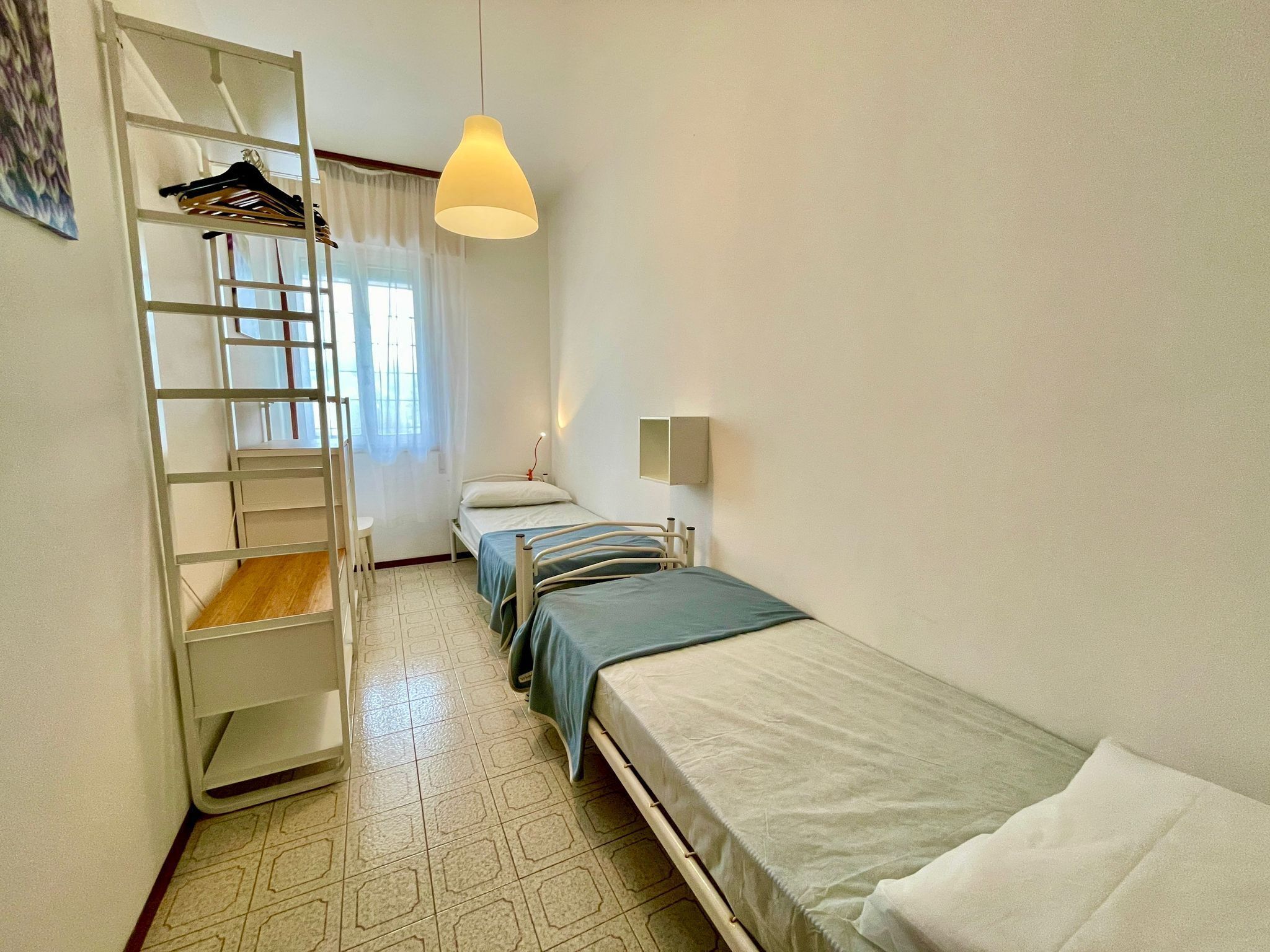 A small, clean room with two single beds, a ladder, a hanging light, and a window with curtains.