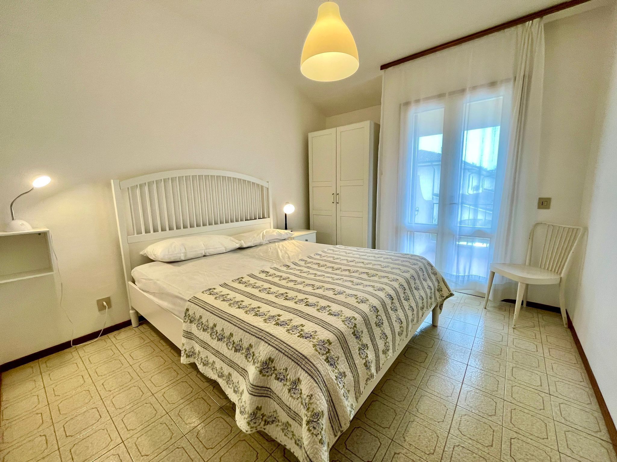 A neatly arranged bedroom with a bed, bedside lamps, a chair, and a window with curtains.
