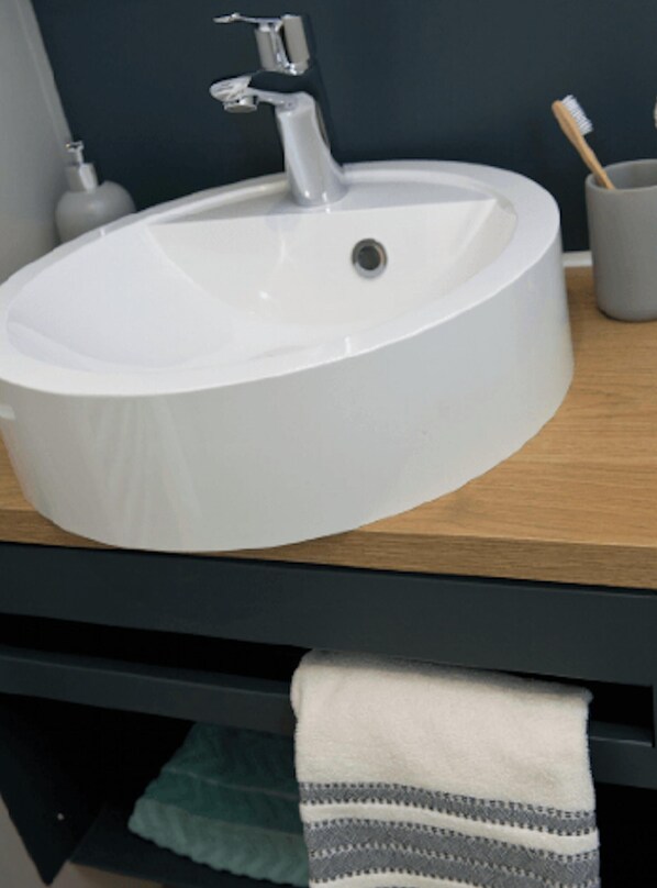 Un lavabo blanc de forme ovale, doté d’un robinet moderne, d’un distributeur de savon et d’un porte-brosses à dents, le tout posé sur un comptoir en bois.