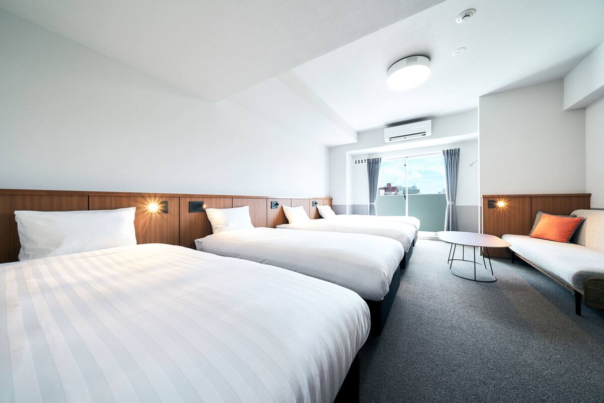 A hotel room with two beds, a sofa, a small table, and a view of the city through a large window.