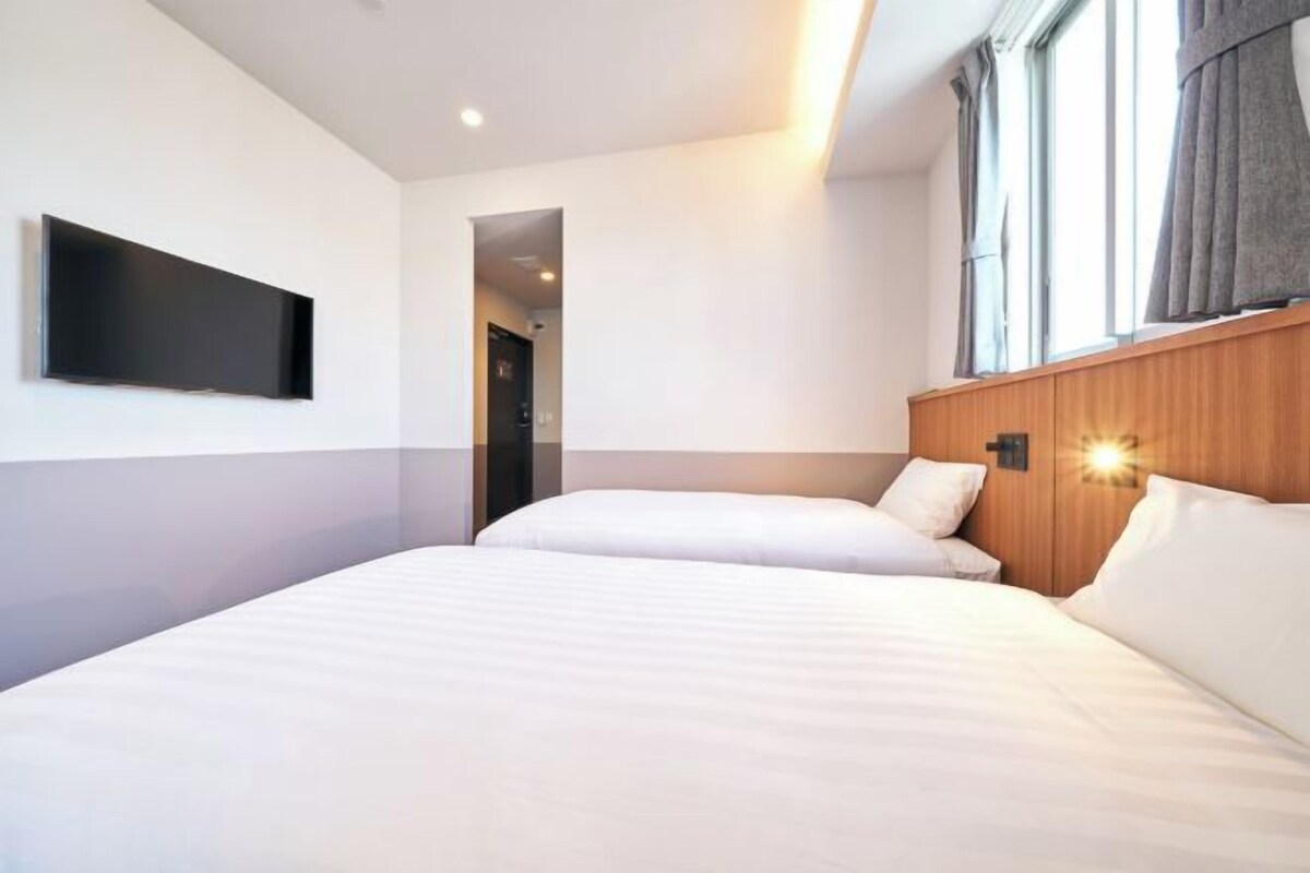 A hotel room with two beds, a flat-screen TV mounted on the wall, and a window with curtains.