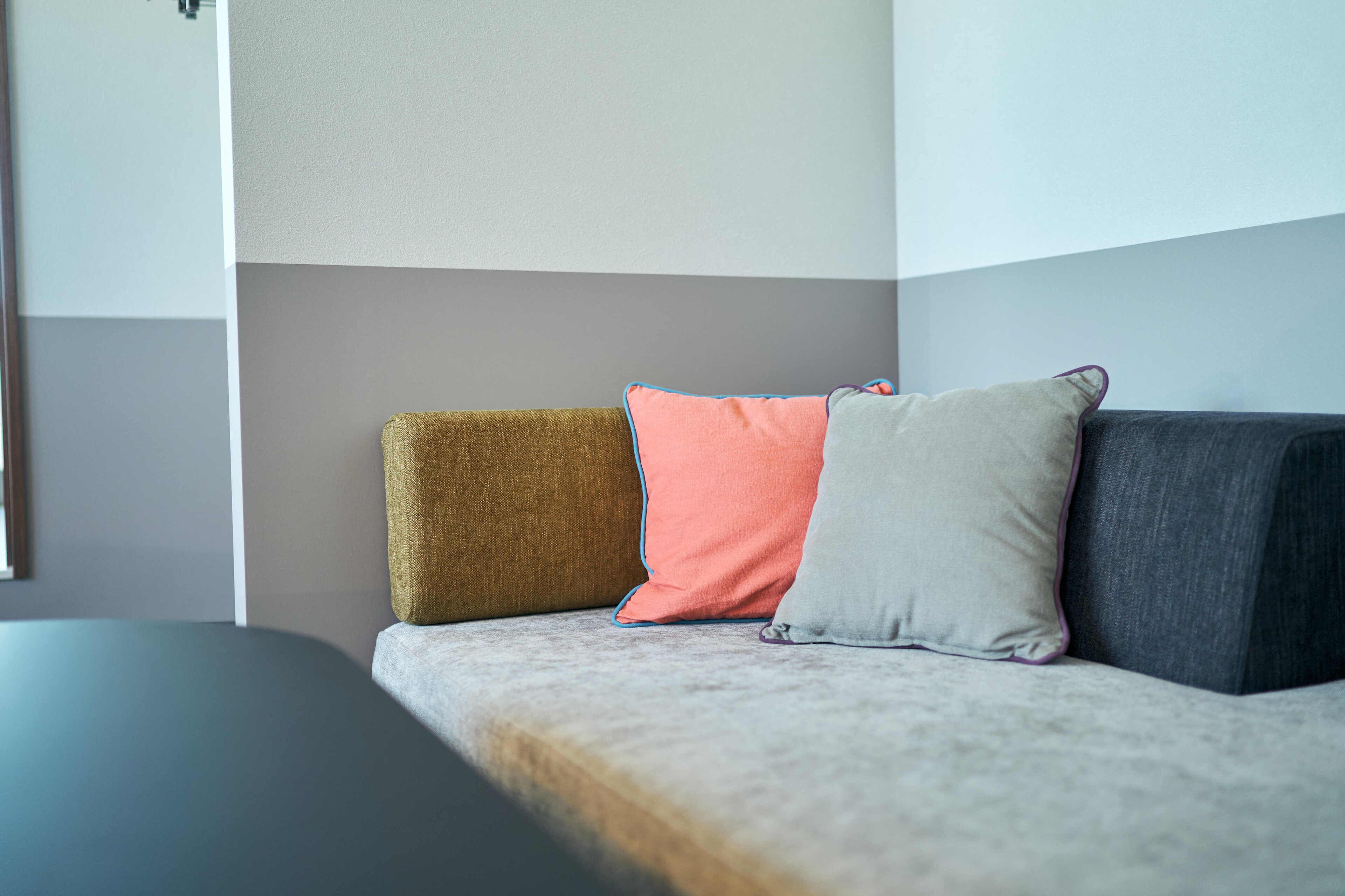 A sofa with three pillows in different colors and a plain wall in the background.