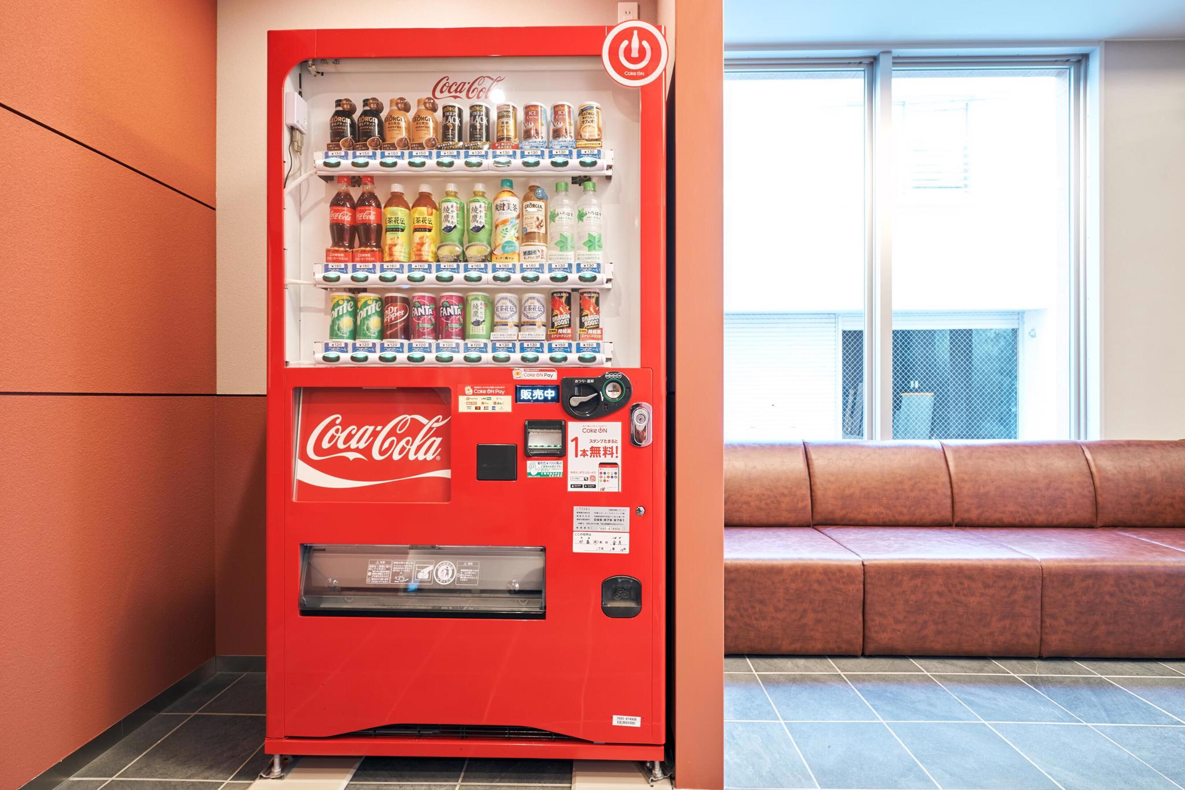 A red Coca-Cola vending machine with various beverages, located next to a window with a view of a building.