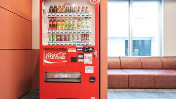 A red Coca-Cola vending machine with various beverages, located next to a window with a view of a building.