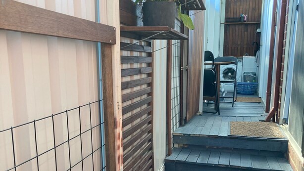 Un estrecho pasillo exterior con pared de azulejos, cerca de madera y un pequeño jardín con césped artificial.