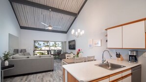 Una cocina moderna con una isla central, un área de comedor con sofá y un amplio ventanal con vistas a palmeras.
