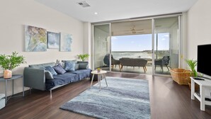 A modern living room with a sofa, a coffee table, and a TV stand. There is a large window with a view of the beach.