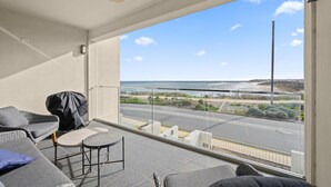 A balcony with a view of the beach and road, furnished with a sofa, chairs, and a table.