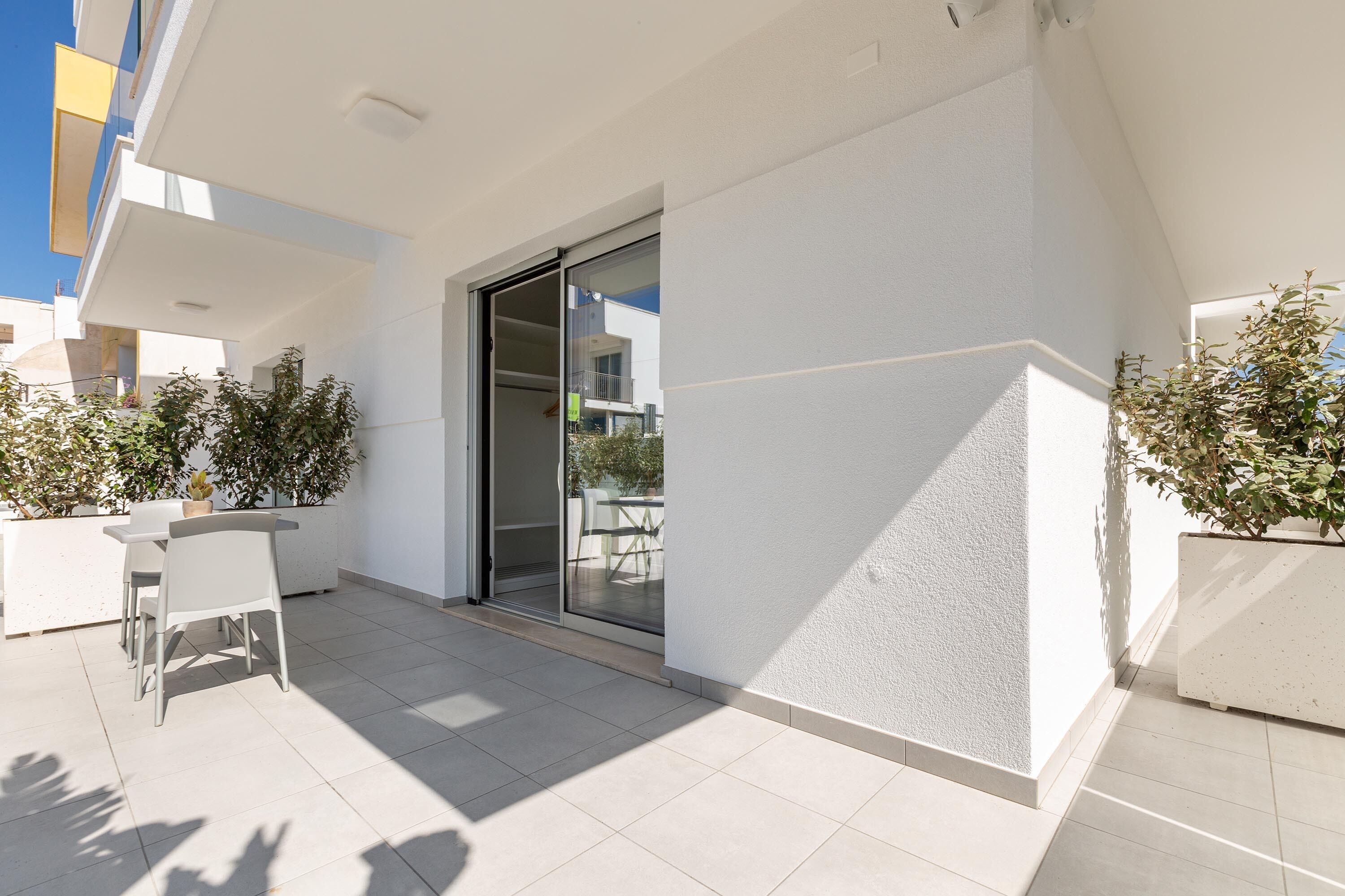 A modern outdoor patio with a sliding glass door leading to a balcony, surrounded by potted plants and a white tiled floor.