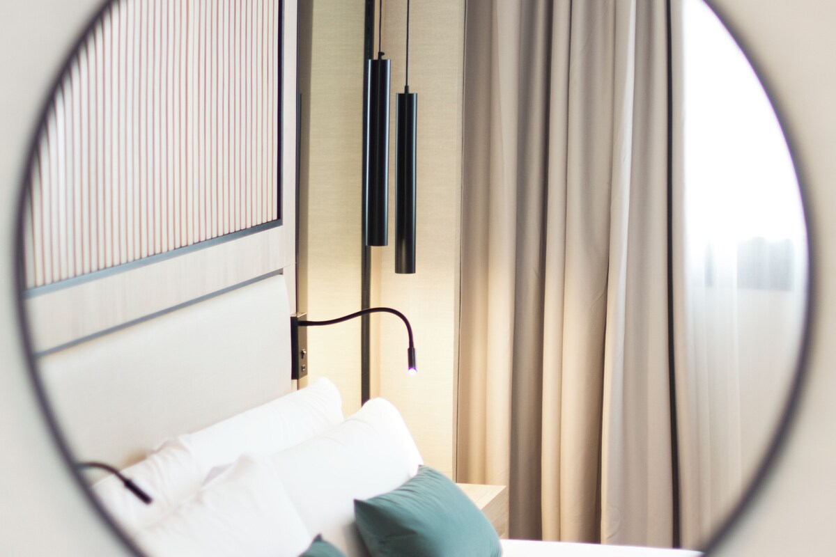 A circular view of a hotel room with a bed, pillows, a nightstand, and curtains.