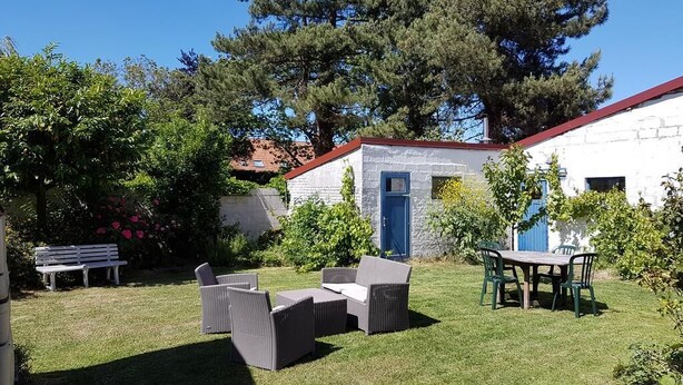A patio area with outdoor furniture, a white building with a blue door, and surrounding greenery.