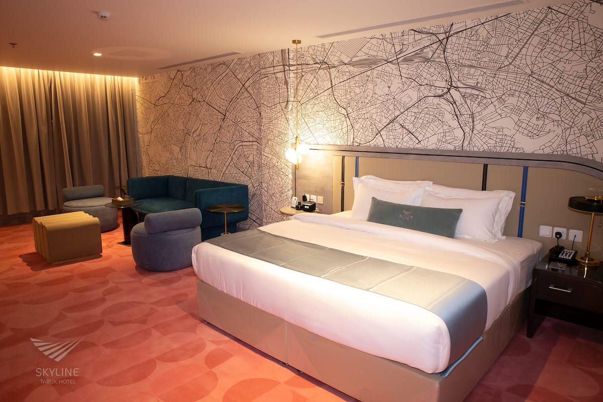 A hotel room with a large bed, a seating area with a sofa and armchairs, a small table, and a wall with a city map design.