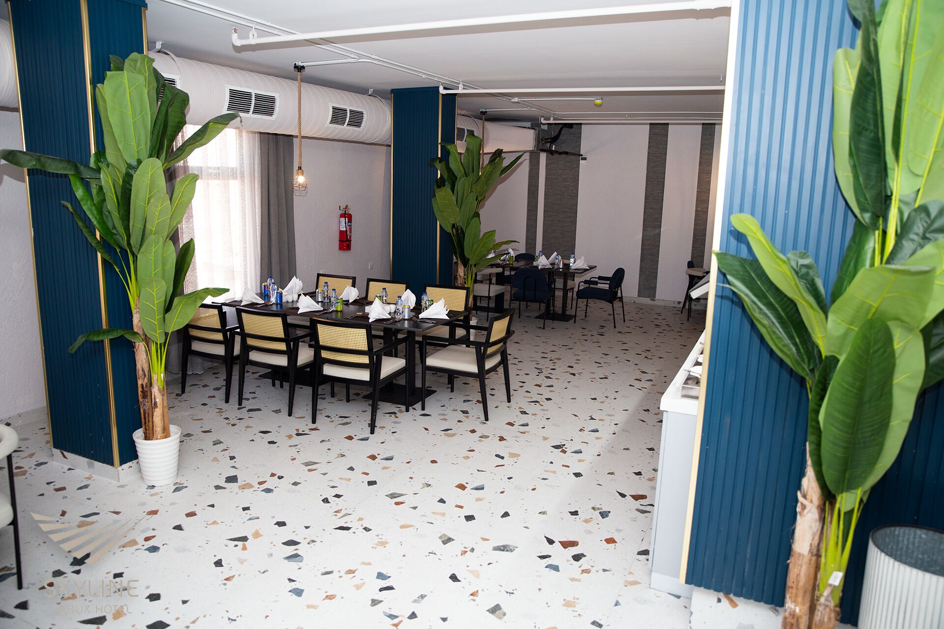 A dining area with a terrazzo floor, wooden tables, and chairs. There are potted plants and a fire extinguisher on the wall.
