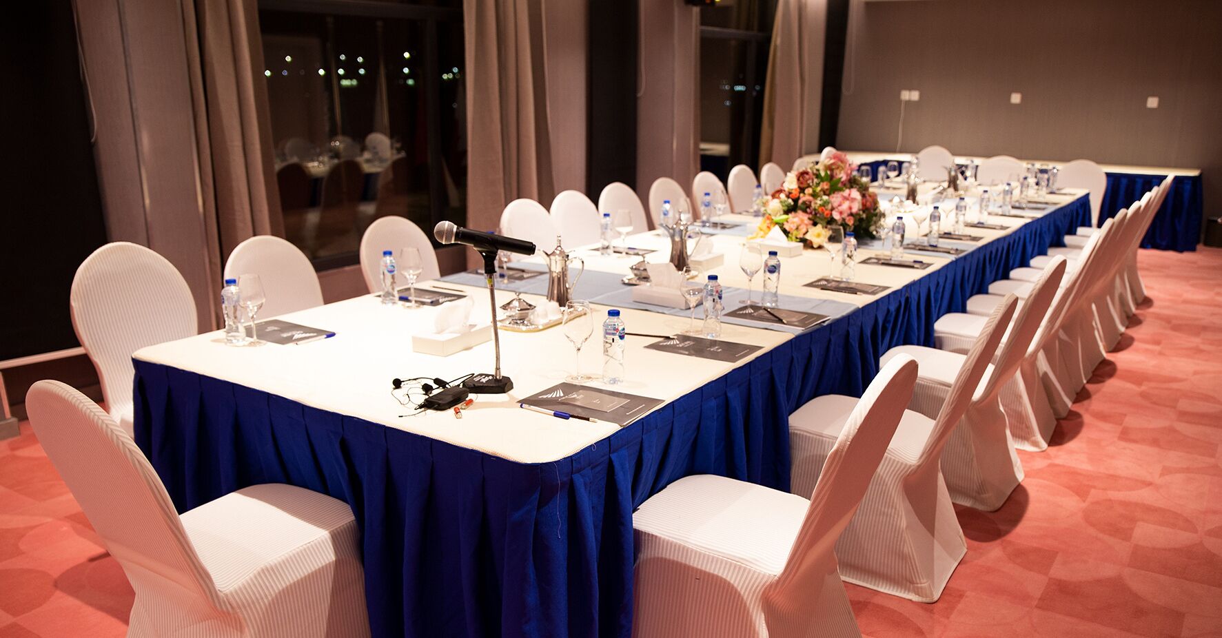 Une salle de conférence avec une longue table, des chaises blanches et un microphone.
