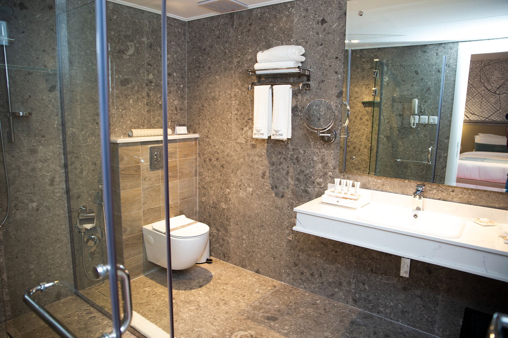 Une salle de bain moderne avec une douche à l’italienne en verre, des toilettes blanches, un grand miroir et un lavabo blanc avec un porte-serviettes au-dessus.