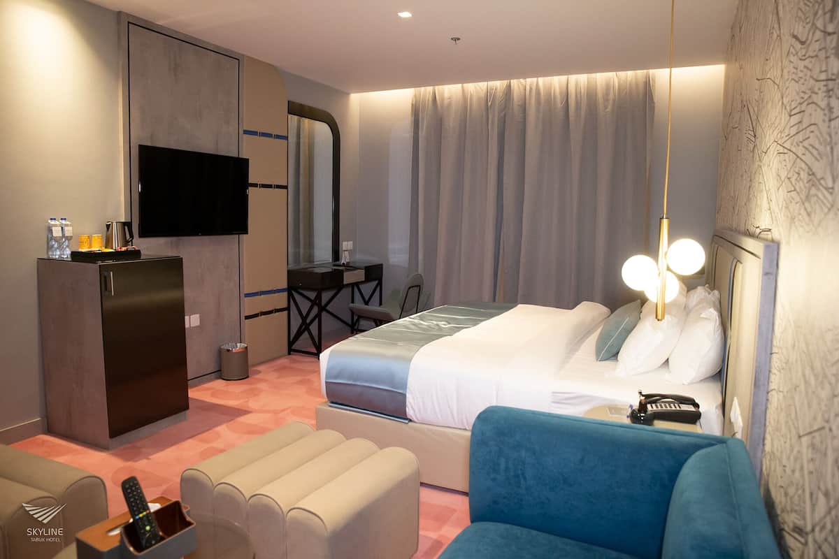 A hotel room with a bed, a desk, a television, a minibar, and a seating area.