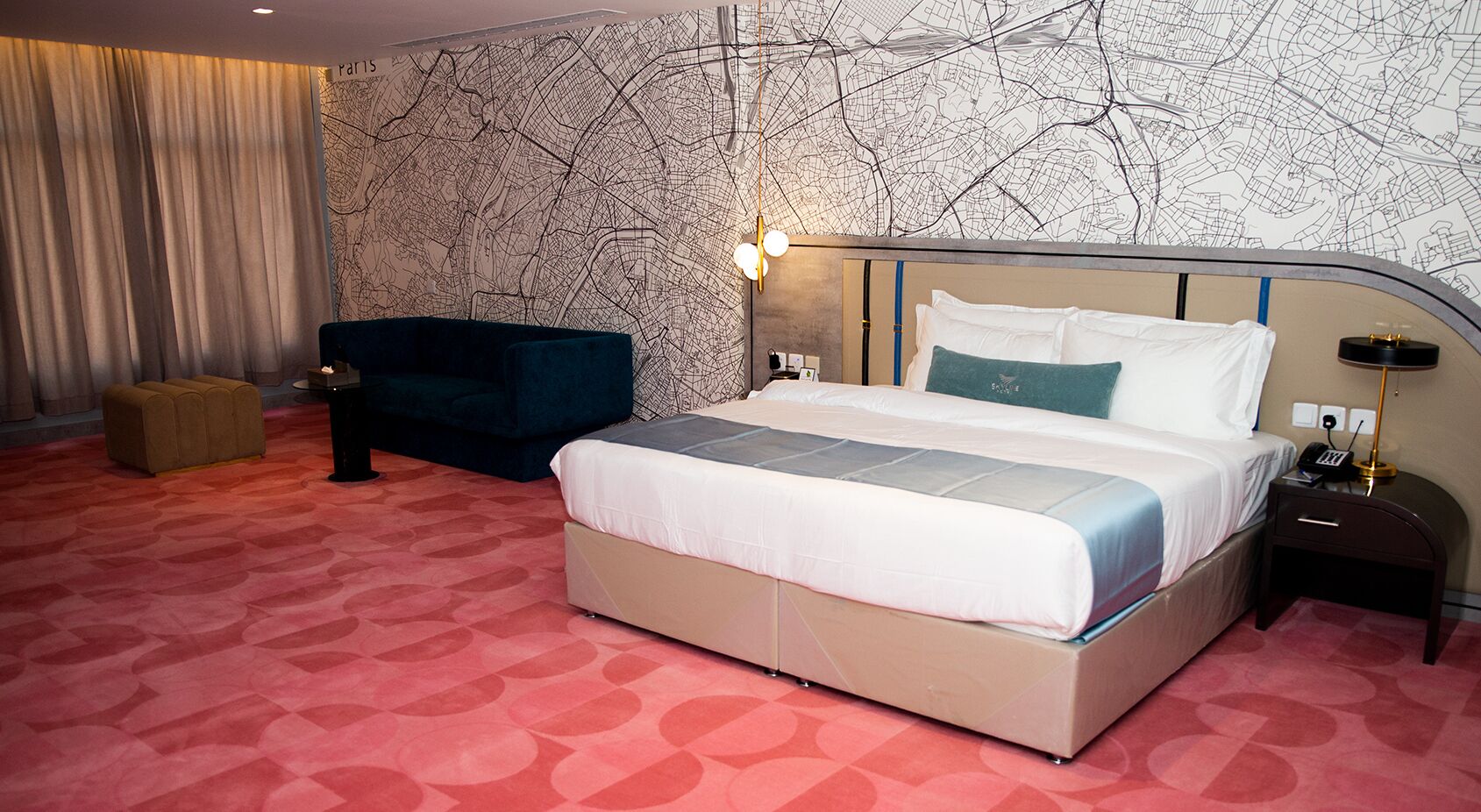 A hotel room with a large bed, a dark blue sofa, a small wooden chest, and a wall with a city map design.