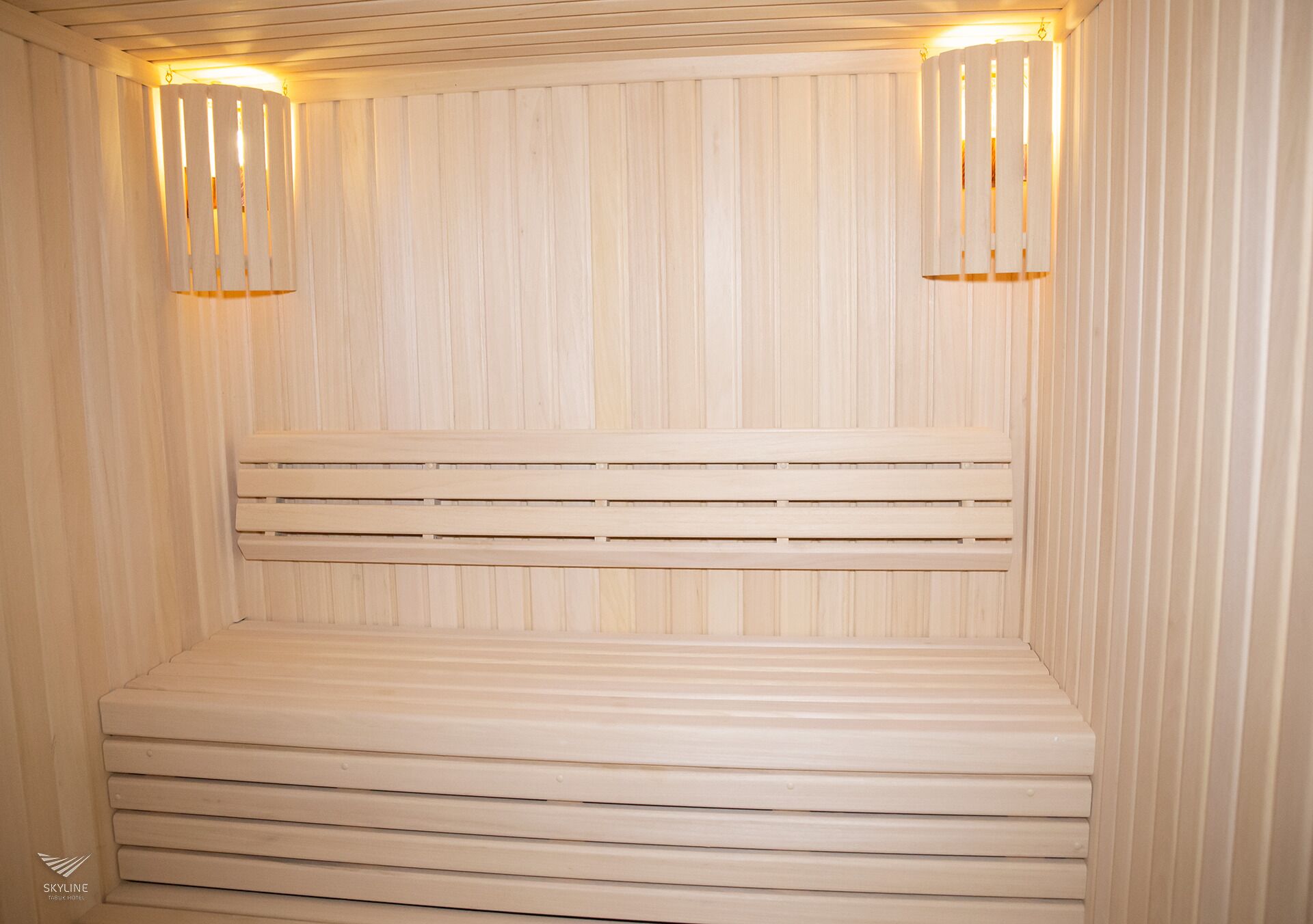 Un sauna doté d’un banc en bois et d’un dossier à lattes, de deux appliques murales et d’un plafond recouvert de panneaux de bois disposés horizontalement.