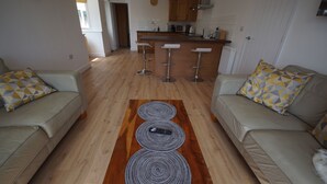 A modern living room with a kitchen in the background, featuring a central island with bar stools, a wooden floor, and a patterned rug. - Vale View - Sleeps 6 - Pets Ok - Garden - Parking (Melton Mowbray)