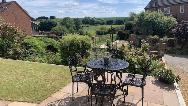 A patio with a round table and four chairs, surrounded by greenery and a view of a rural landscape. - Vale View - Sleeps 6 - Pets Ok - Garden - Parking (Melton Mowbray)