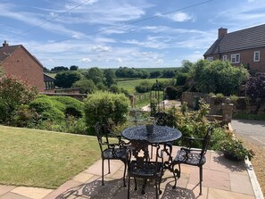 A patio with a round table and four chairs, surrounded by greenery and a view of a rural landscape. - Vale View - Sleeps 6 - Pets Ok - Garden - Parking (Melton Mowbray)