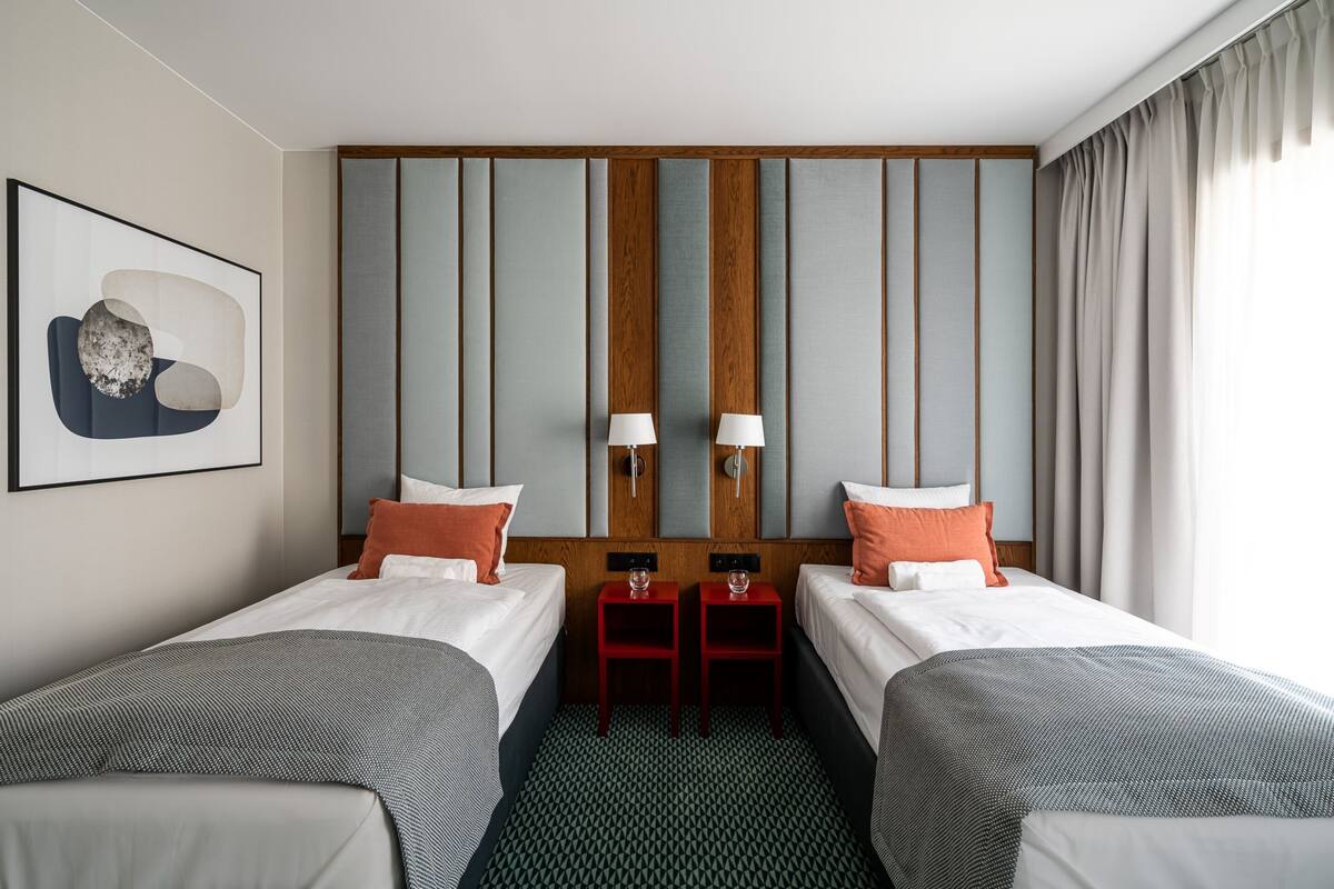 Ein Hotelzimmer mit zwei Betten, einem Holzkopfteil, zwei Nachttischen mit Lampen, einem gerahmten Kunstwerk an der Wand und einem gemusterten Teppich.