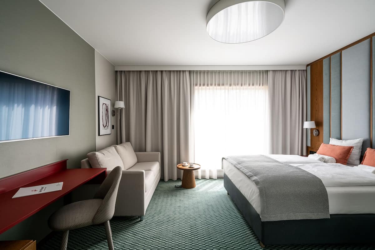 Ein Hotelzimmer mit Bett, Schreibtisch, Stuhl, kleinem Tisch und einem großen Fenster mit Vorhängen.
