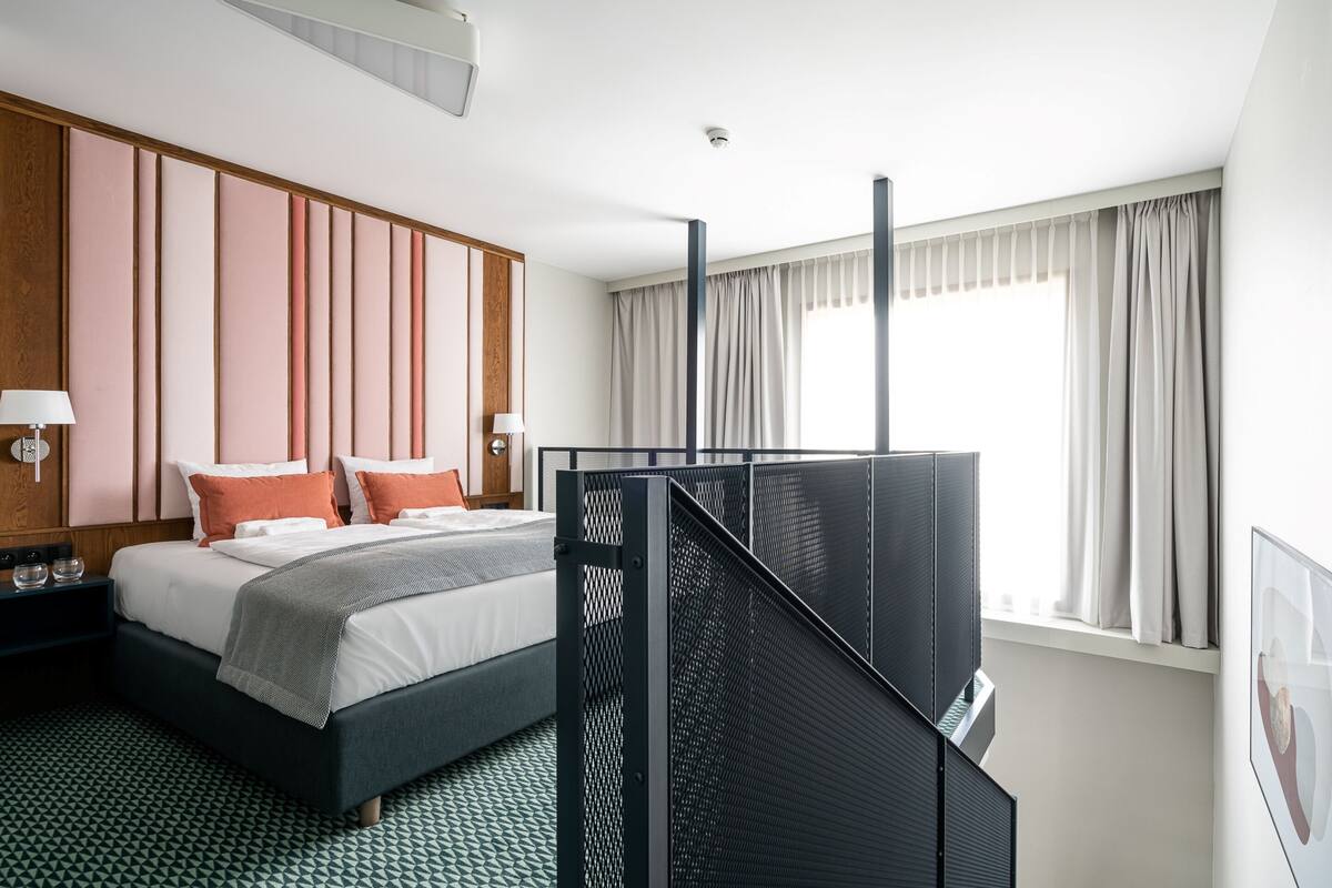 Ein Hotelzimmer mit Bett, Schreibtisch, Stuhl, einem Fenster mit Vorhängen und einem an der Wand befestigten Fernseher.