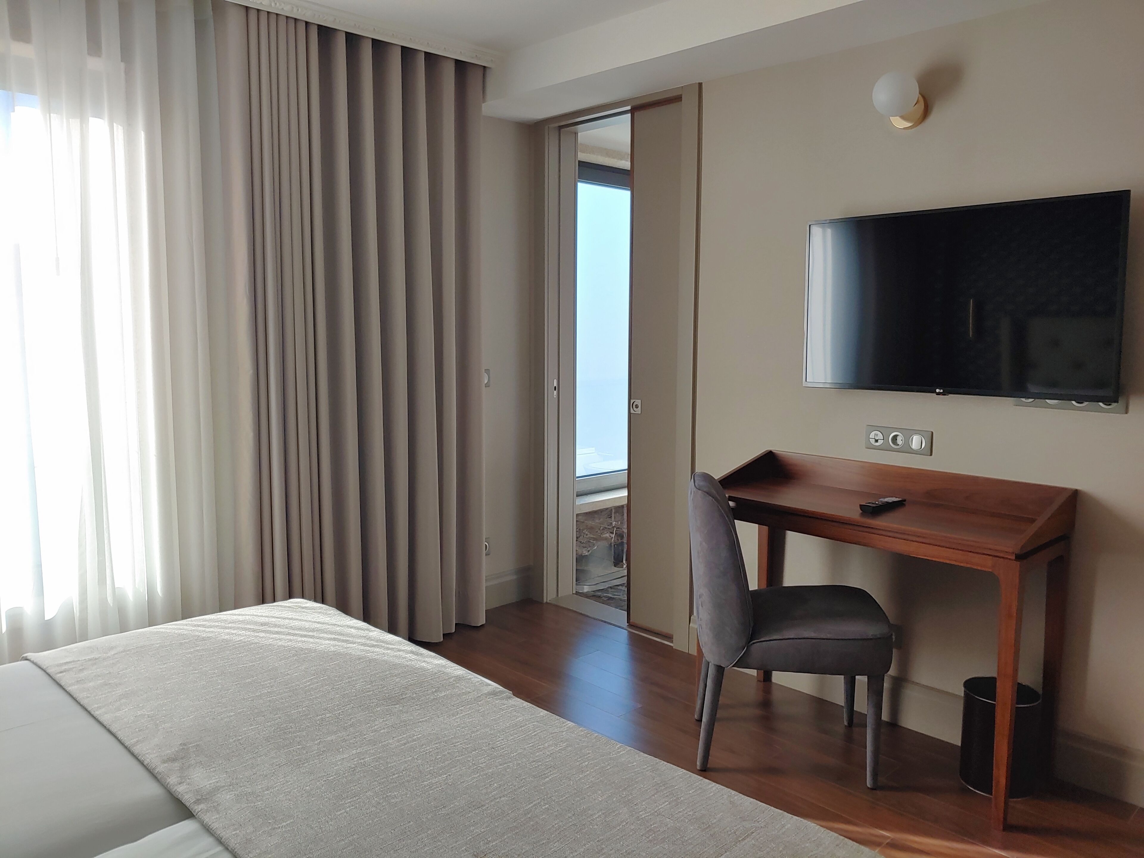 A hotel room with a bed, a desk, a chair, a TV, and a view through a door.