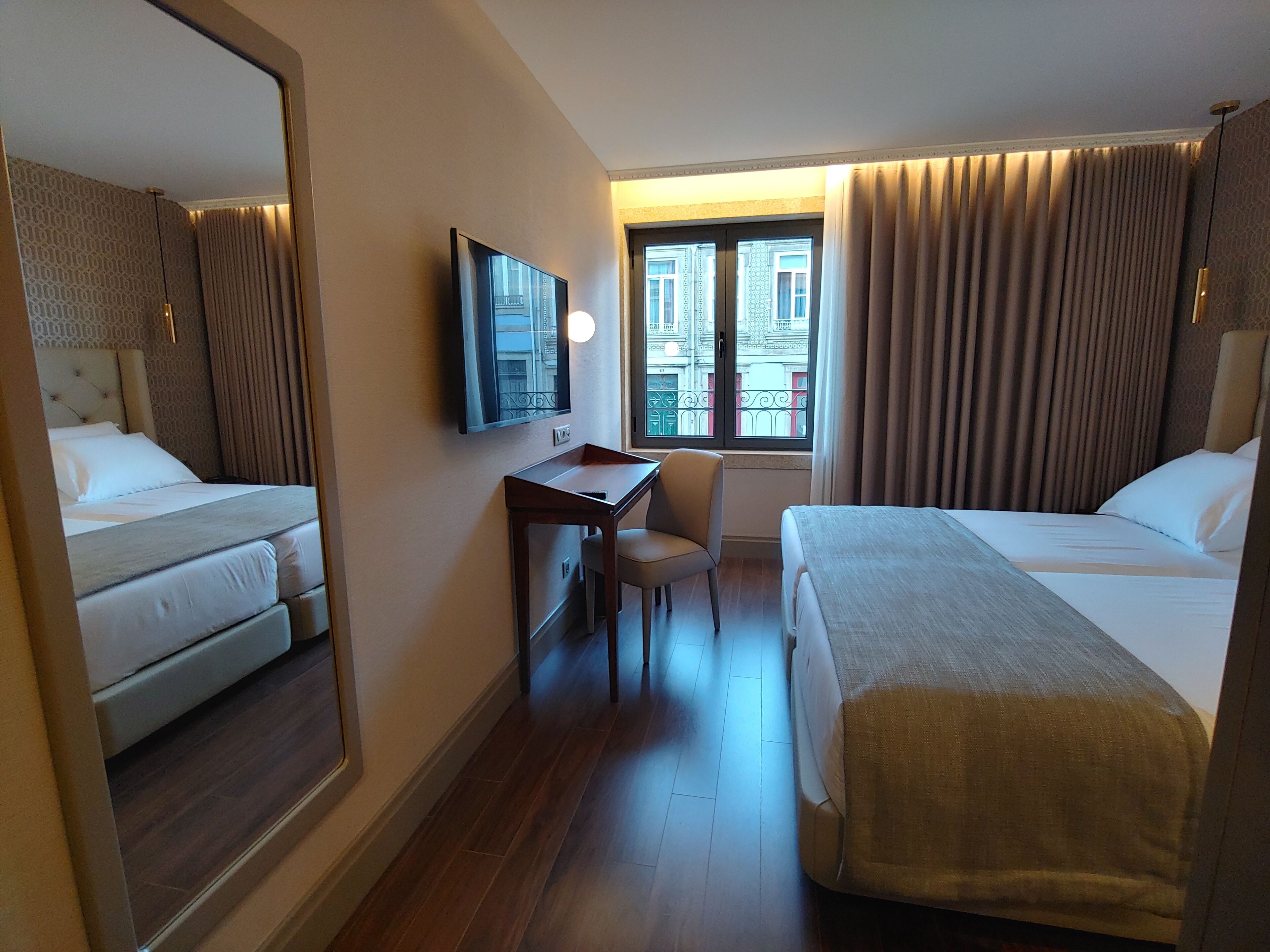 A hotel room with two beds, a desk, a chair, a TV, and a window with curtains.