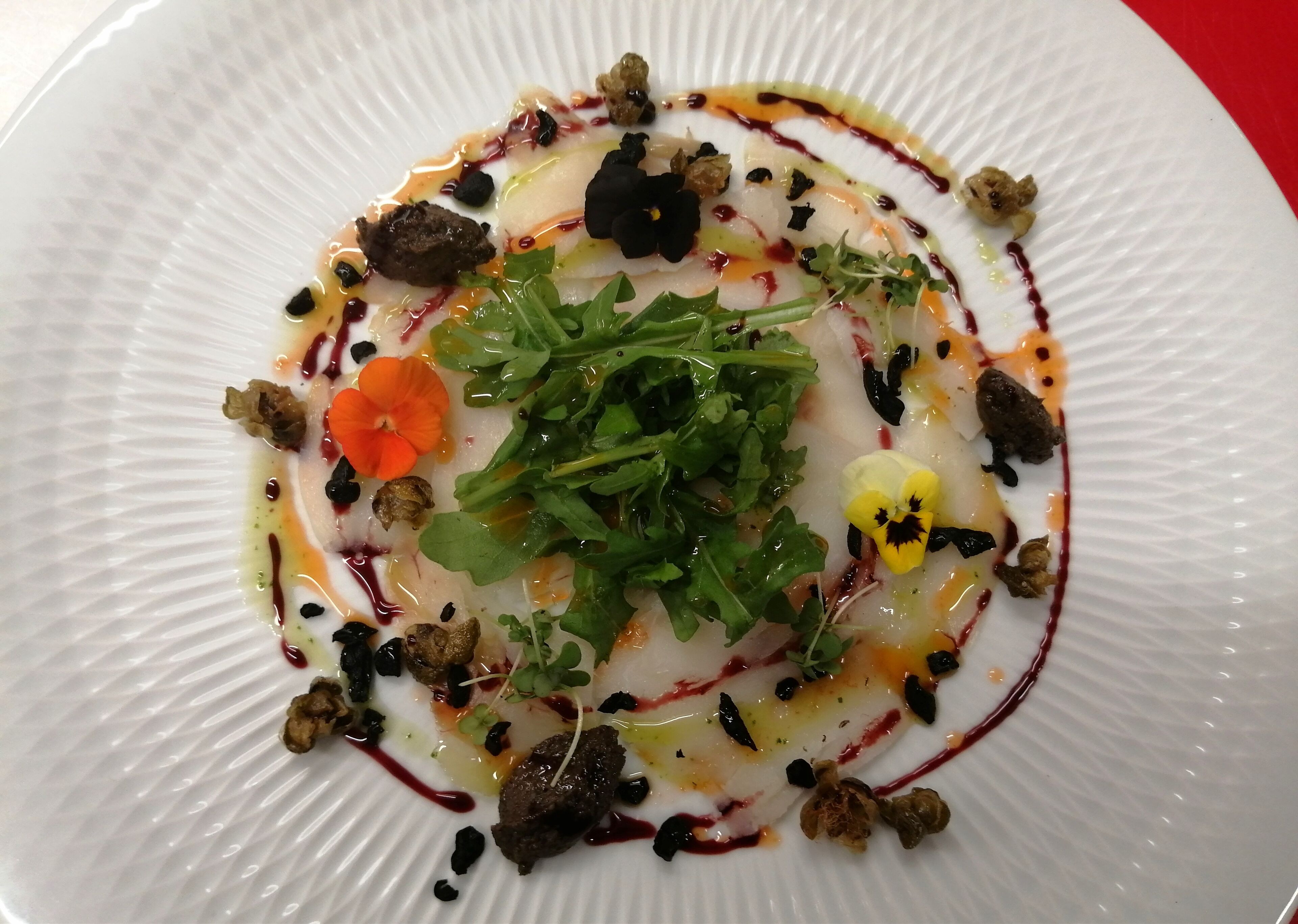 A white plate with a dish of food, including greens, a flower, and some dark elements.