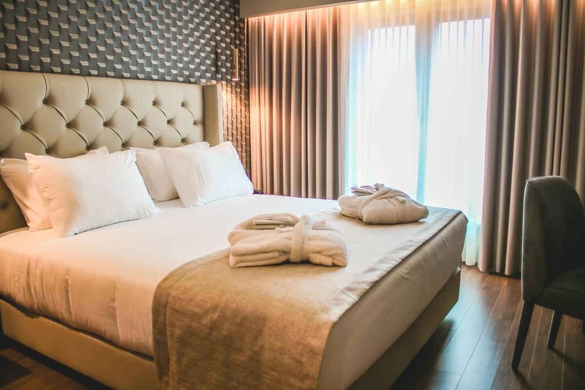 Een net opgemaakt bed met witte lakens, een hoofdbord met een geometrisch patroon, en een paar handdoeken op het bed.