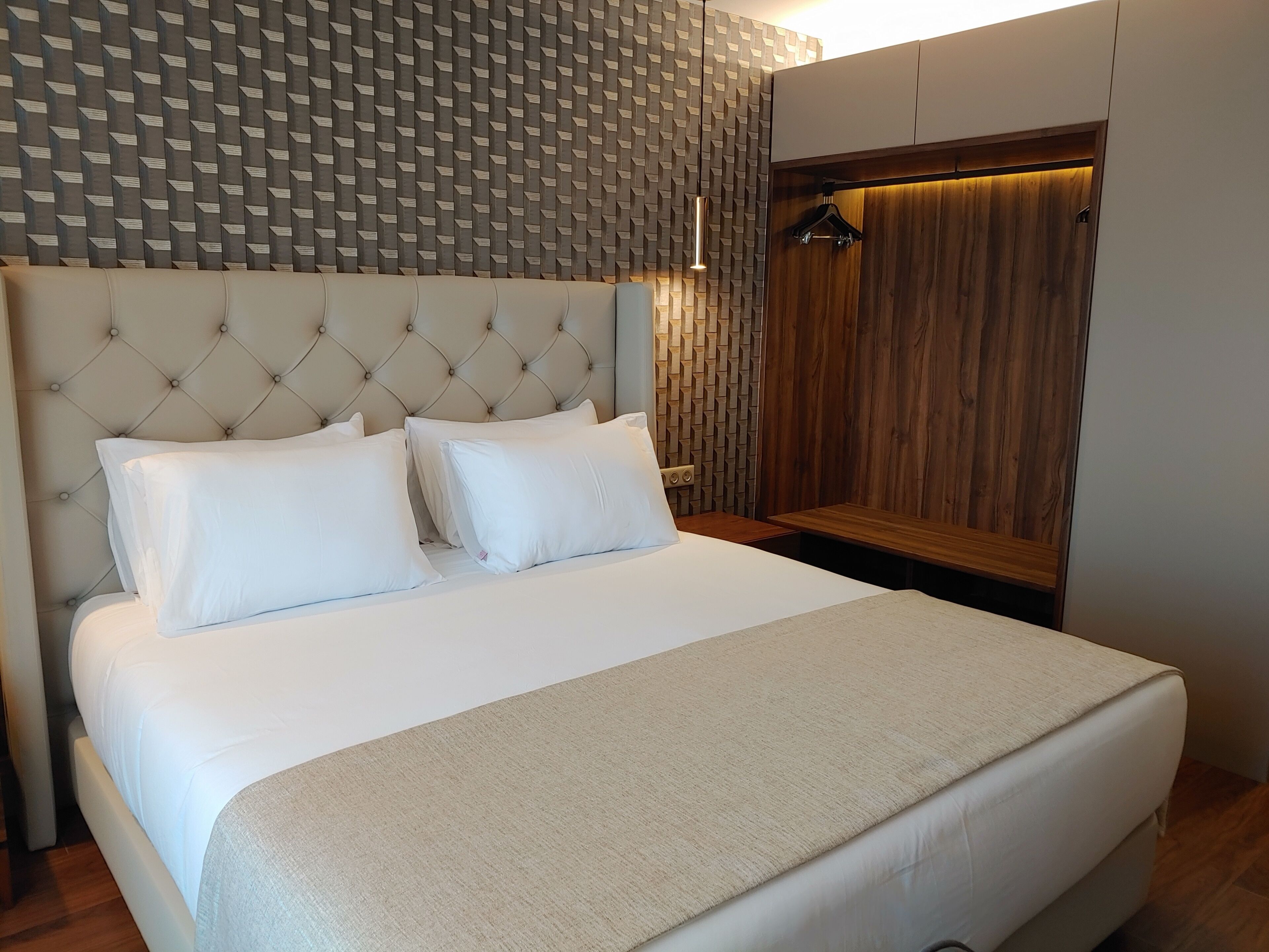 Una cama bien tendida con sábanas blancas y un cabecero capitonado, un banco de madera y una pared decorada.