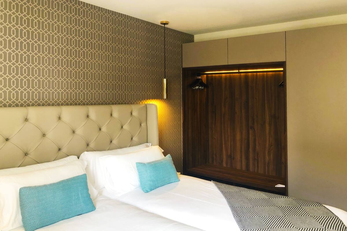 Een hotelkamer met een gestoffeerd hoofdeinde, een kamer met een patroon, een houten garderobe en een bed met witte en blauwe kussens.