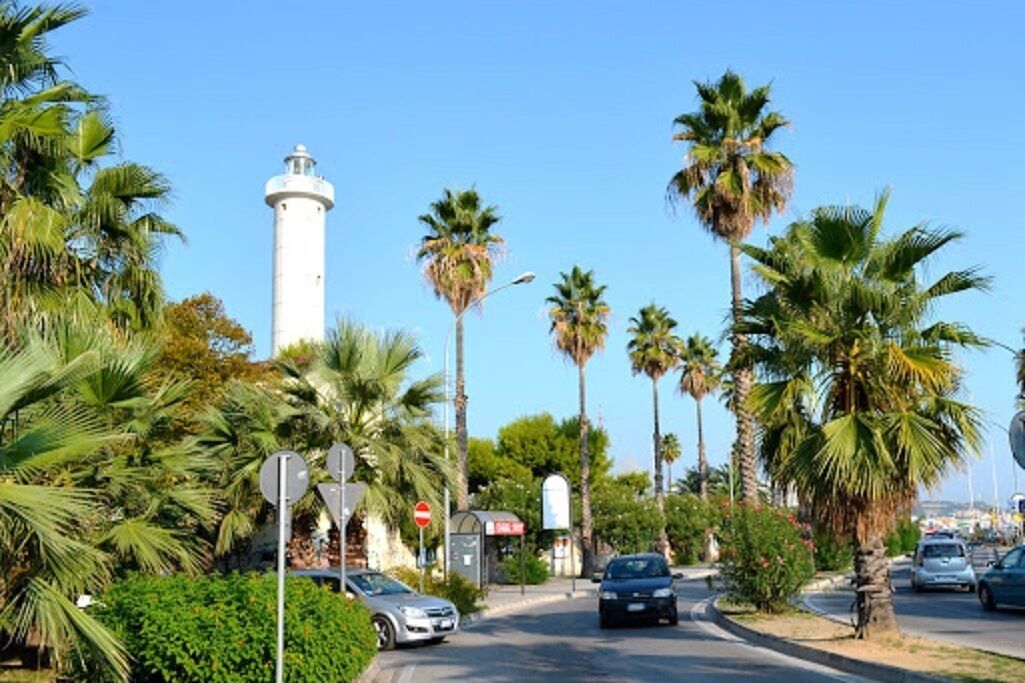 A street with palm trees, a lighthouse, and parked cars.