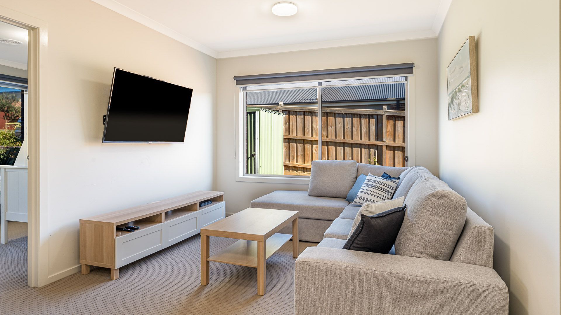 Ein modernes Wohnzimmer mit einem an der Wand befestigten Flachbildfernseher, einem Holztisch, einem beige farbenen Sofa mit Kissen und einem großen Fenster mit Blick auf einen Holzzaun.