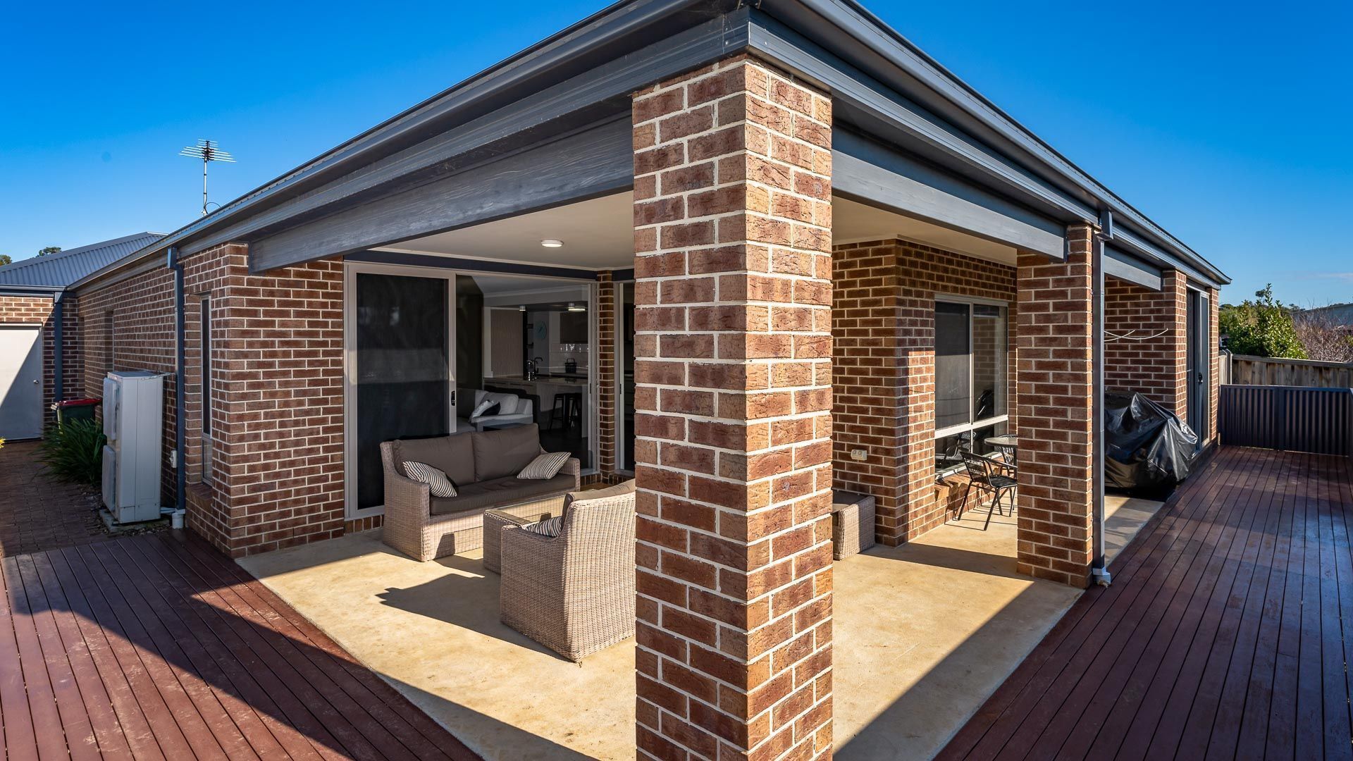 A covered patio area with a brick exterior, a sliding glass door leading to an indoor space, and a wooden deck.