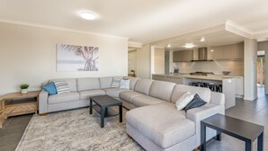 A modern living room with a sectional sofa, a coffee table, and a rug. The room has a kitchen in the background with wooden cabinets and a countertop.