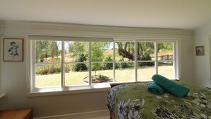 A bedroom with a large window, a bed with a floral bedspread, a wooden nightstand, and a framed picture on the wall. - Bylands Cottage. Historic home with Tambo River views (Swan Reach)