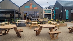 An outdoor seating area with wooden tables and chairs, a building with signs for 'Drink Relax Dance' and 'RECEPTION', and a 'Welcome' sign. - St Osyth- Sleeps8-freeparking-wifi-holidaypark (Clacton-on-Sea)