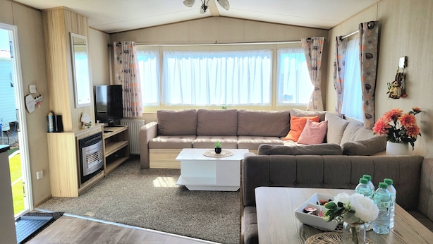 A cozy living room with a sofa, coffee table, and a TV. There is a fireplace, a window with curtains, and a ceiling fan. - St Osyth- Sleeps8-freeparking-wifi-holidaypark (Clacton-on-Sea)