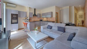 Un saló modern amb un sofà seccional gris, una taula de centre blanca i una zona de cuina amb armaris grisos i un rebedor de maó.