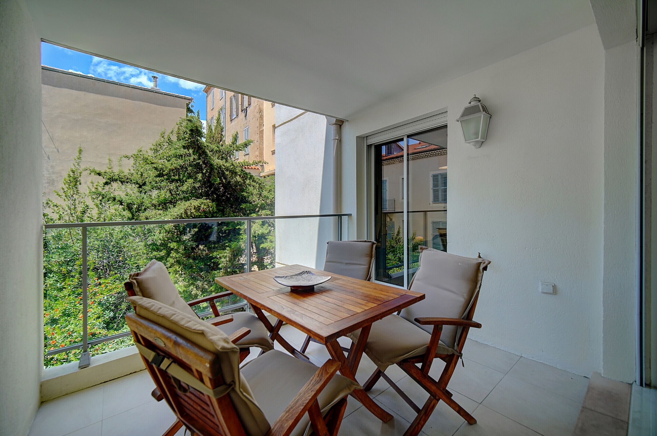 A balcony with a wooden table and chairs, a view of greenery and buildings, and a white wall with a window and a light fixture.