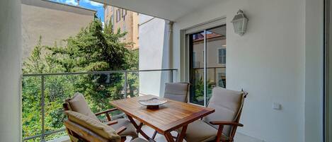 A balcony with a wooden table and chairs, a view of greenery and buildings, and a white wall with a window and a light fixture.