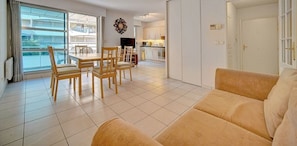 A living room with a sofa, a dining table with chairs, a TV, and a kitchen in the background.