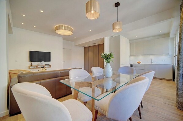 A modern living room with a glass dining table, white chairs, a brown sofa, and pendant lights.