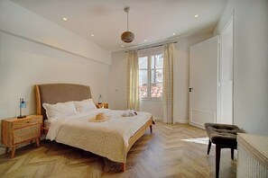 Quarto com cama grande, mesas de cabeceira, uma cadeira, janela com cortinas e uma porta.
