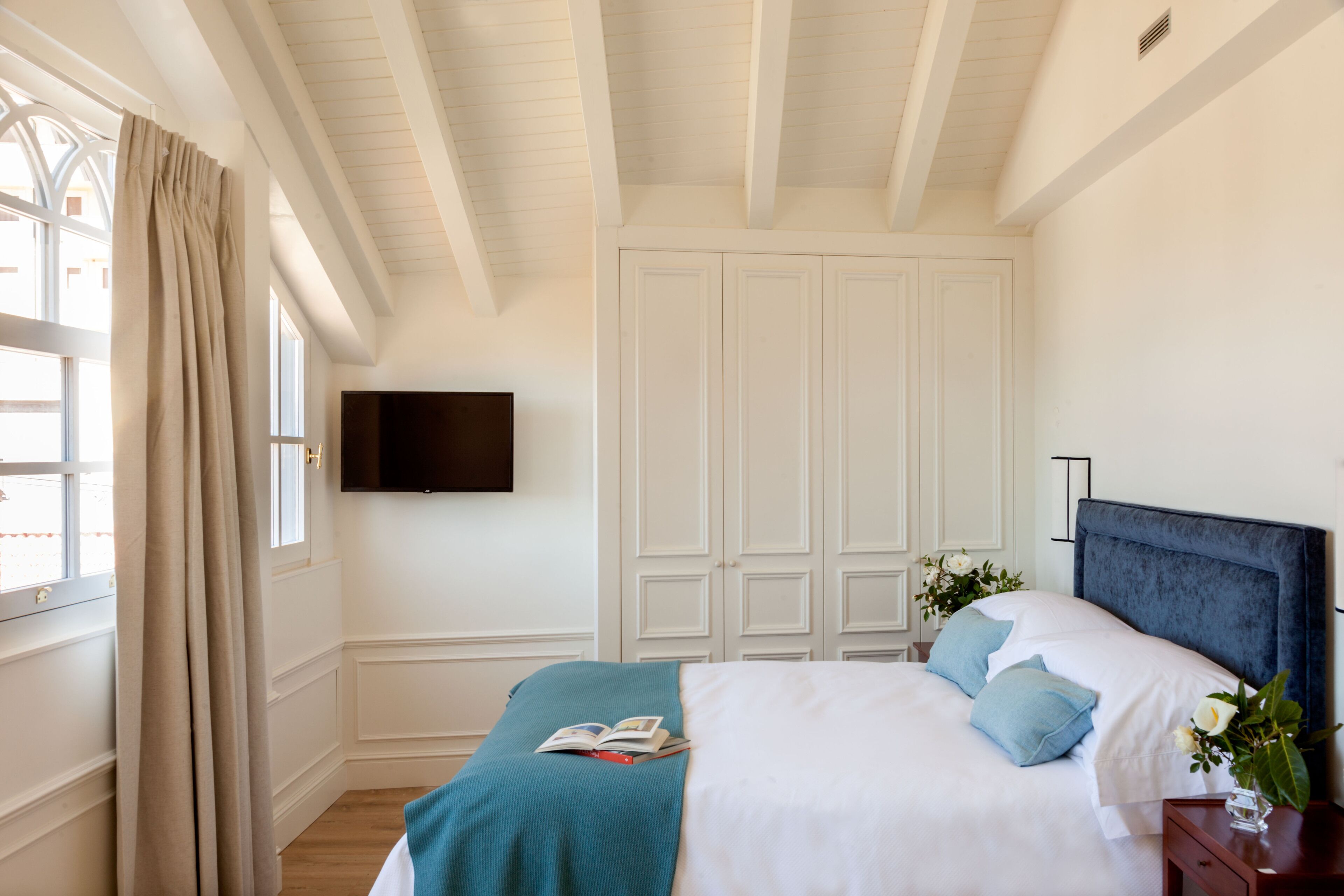 A bedroom with a bed, a TV mounted on the wall, a window with curtains, and a wardrobe.