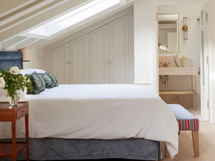 A bedroom with a skylight, a bed, a bedside table with a vase of flowers, a bench, and a bathroom with a sink and mirror.