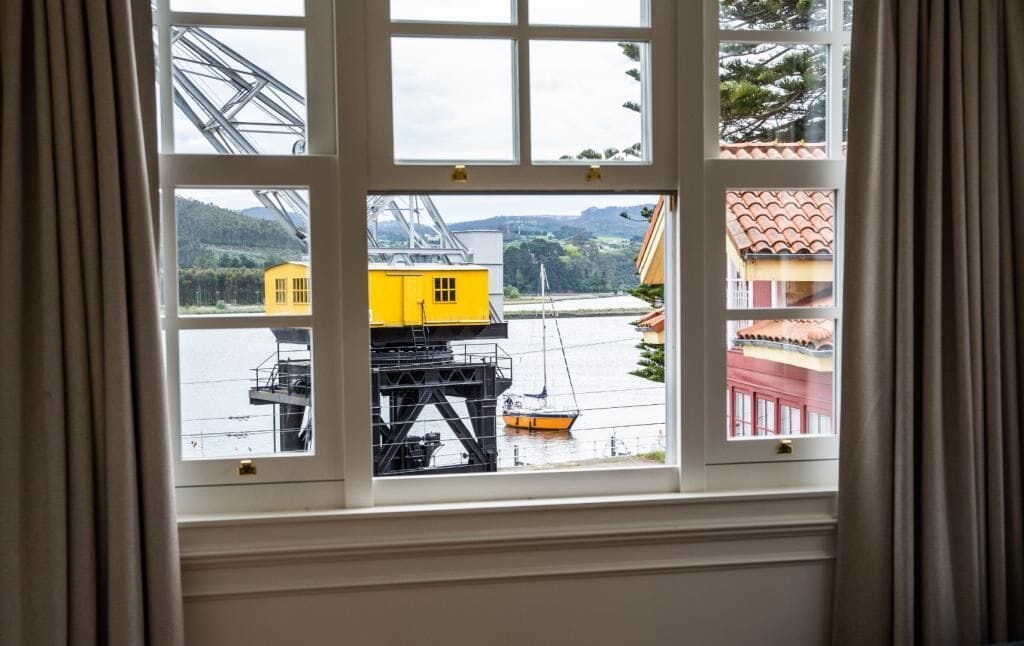 A window with a view of a yellow crane and a sailboat on a body of water, with buildings in the background.